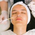 pexels-photo-5069432-5069432 Top view skilled female cosmetologist in uniform and gloves applying essential oil on young woman face skin while working in contemporary spa center